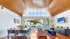 Uma sala de estar ampla com piso de madeira, uma mesa de jantar com cadeiras, um sofá e uma TV fixada na parede.