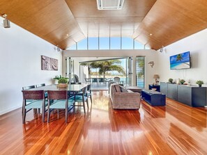 A spacious living area with a wooden floor, a dining table with chairs, a sofa, and a TV mounted on the wall. - Solmar At The Waters Edge (Nelly Bay)