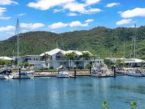 A marina with boats docked, a large house with a white roof, and a forested hill in the background. - Solmar At The Waters Edge (Nelly Bay)