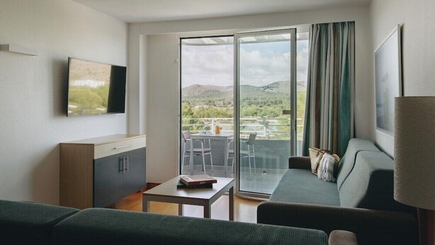 Ett modernt hotellrum med en soffa, ett litet bord, en TV som är monterad på väggen och en balkong med utsikt över landskapet.