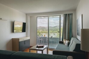 A modern hotel room with a sofa, a small table, a TV mounted on the wall, and a balcony with a view of the countryside. - Ivory Playa (Alcúdia)