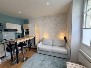 A compact living space with a kitchenette, a sofa, a dining table with chairs, and a patterned wall. - Petit Studio au coeur de Biarritz (BIARRITZ)