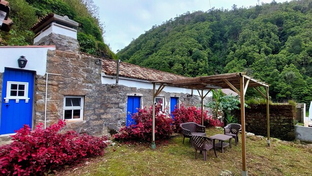 Un edificio de piedra con puertas y ventanas azules, rodeado de abundante vegetación y una colina boscosa.