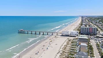 A beachside view with a pier extending into the ocean, beachfront properties, and a road parallel to the beach.