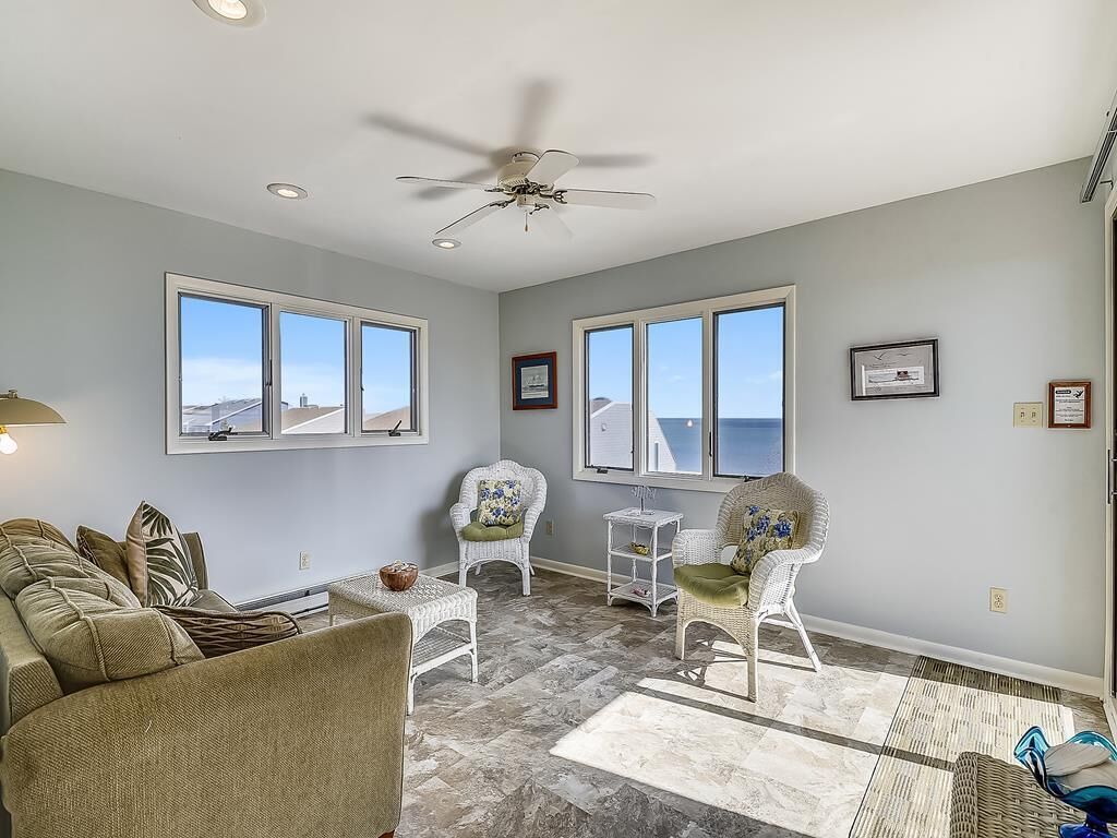 A spacious living room with a ceiling fan, large windows, a sofa, two chairs, and a small table.