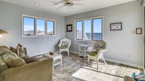 A spacious living room with a ceiling fan, large windows, a sofa, two chairs, and a small table. - Walk to Beach | 4BR Beach House w/ Decks + Kitchen (Tower Shores)