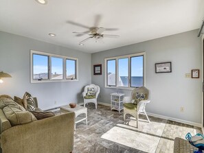 A spacious living room with a ceiling fan, large windows, a sofa, two chairs, and a small table. - Walk to Beach | 4BR Beach House w/ Decks + Kitchen (Tower Shores)