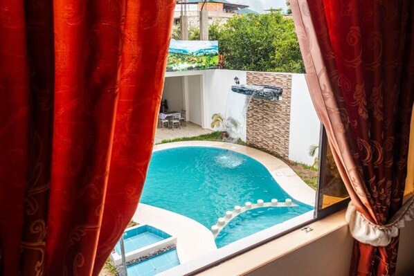 A view of a swimming pool with a waterfall feature, surrounded by a stone wall and greenery, visible through red curtains.
