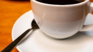 Una taza de café sobre un platito con una cuchara, sobre una mesa con un plato de pasteles al fondo.