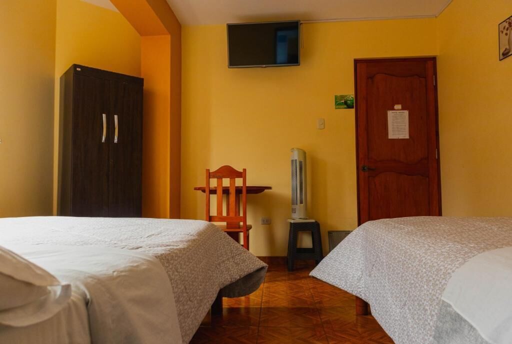 Habitación de hotel con dos camas, un televisor montado en la pared, una puerta de madera y una mesita con una silla.