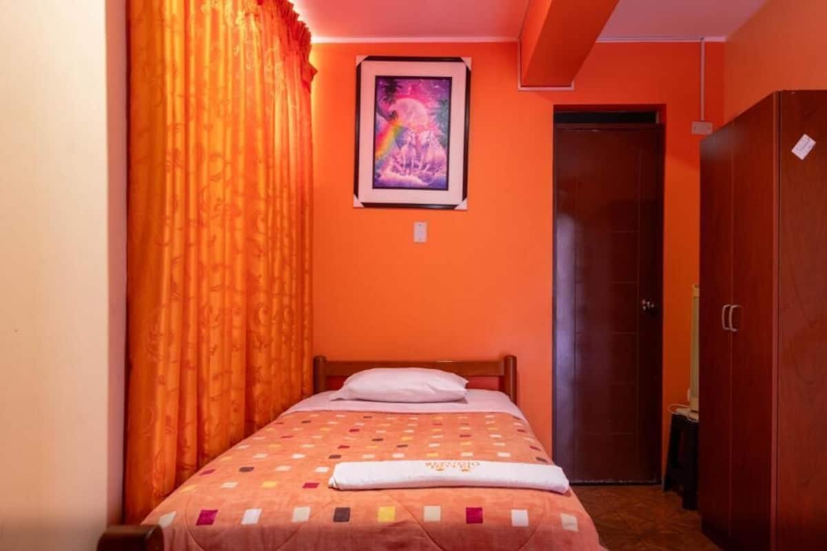 A single bed with a colorful quilt, an orange wall, a framed picture, and a wooden wardrobe.