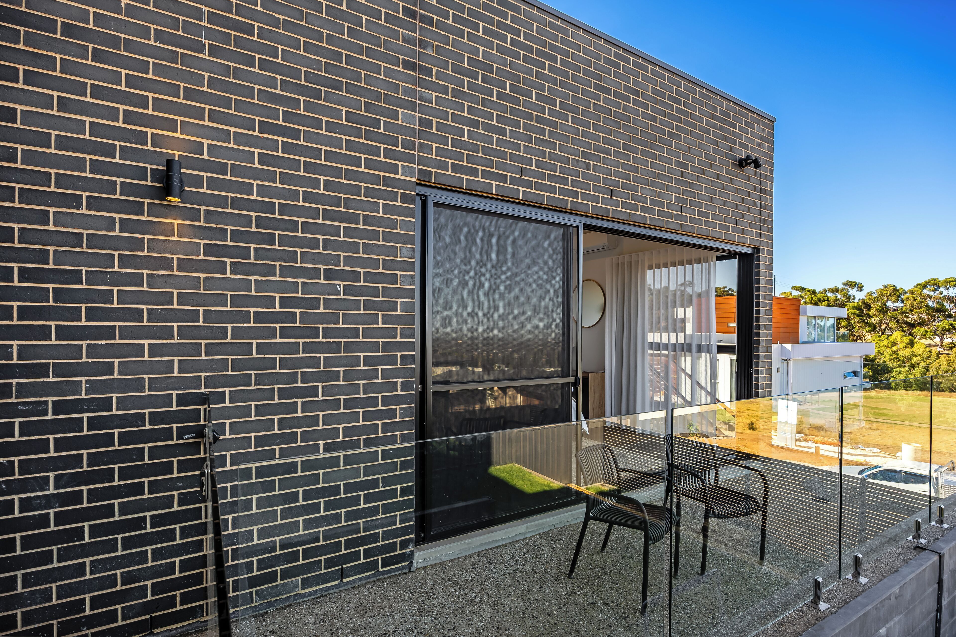 A modern building with a brick facade, large sliding glass doors, and a glass railing overlooking a landscaped area.