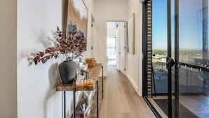 Un couloir moderne avec un plancher en bois, une porte en verre à cadre noir donnant sur un balcon, et un vase contenant des feuilles séchées, posé sur une console en bois.