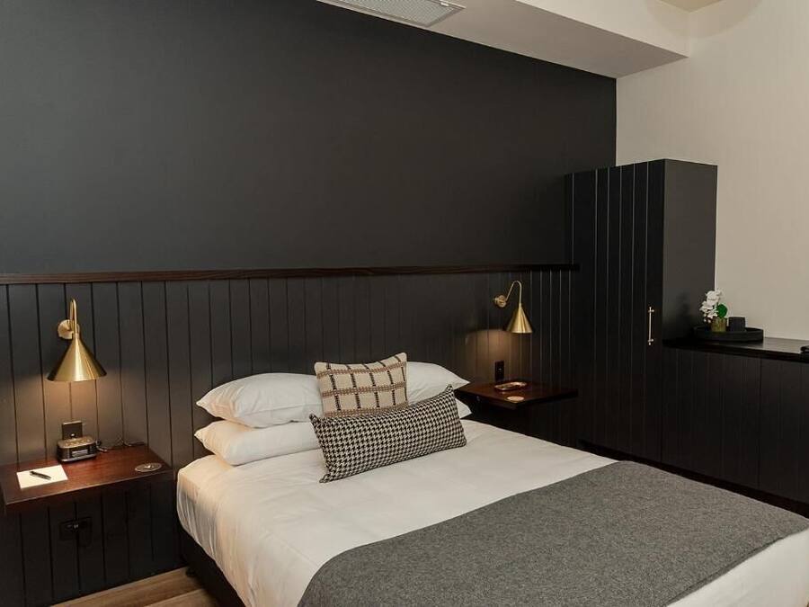 Un dormitorio moderno con cabecera de madera oscura, una cama con ropa de cama blanca, dos lámparas de noche y una mesita de noche con lámpara.