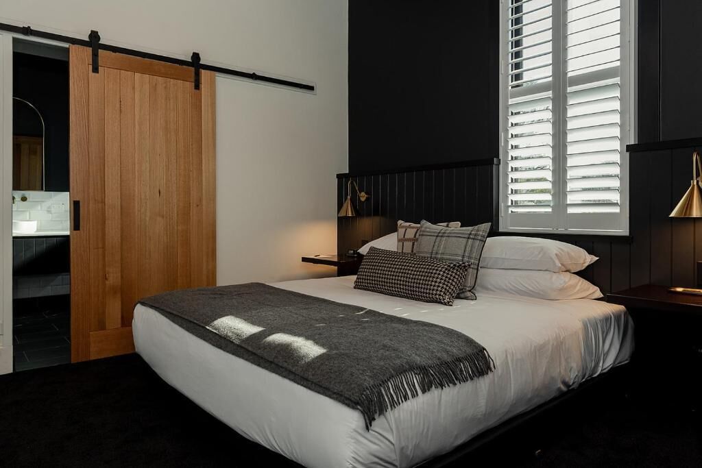 A bedroom with a large bed, a wooden sliding door, a window with blinds, and a bedside table with a lamp.