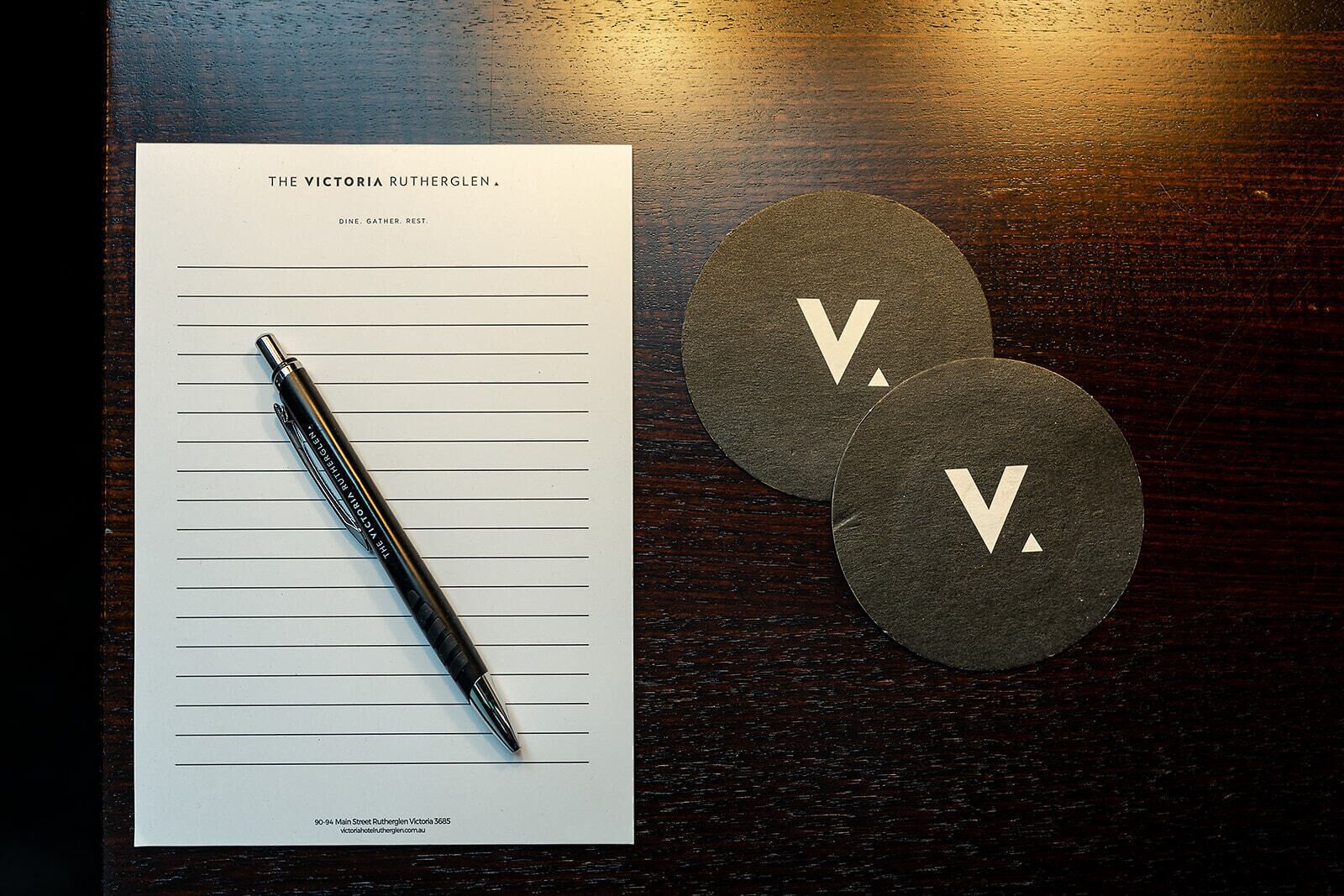 Een notitieboekje met de titel 'The Victoria Rutherglen', met een pen erop, en twee ronde onderzetters met de letter 'V' op een houten oppervlak.