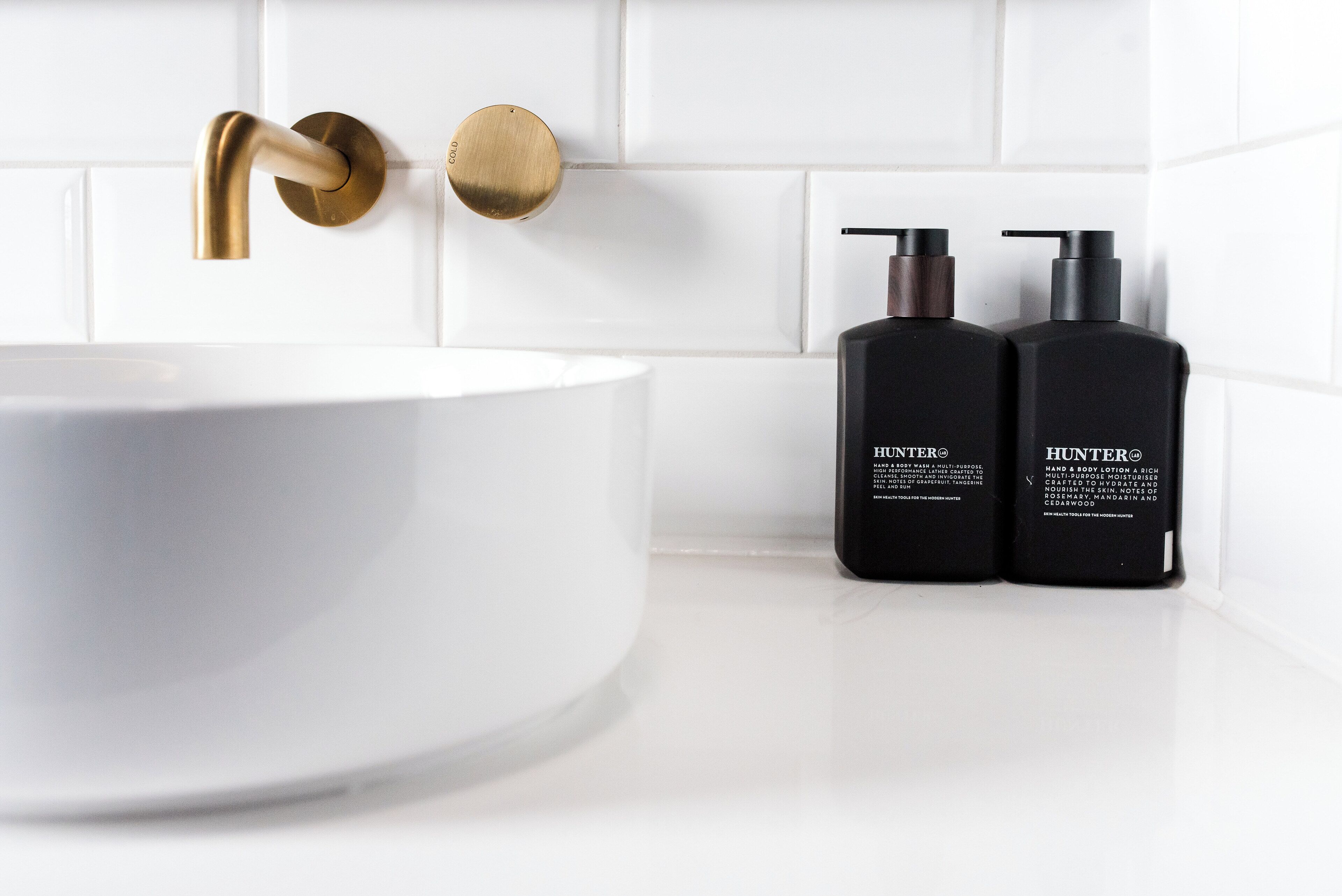 A bathroom with a white sink, gold faucet, and two black Hunter hand soap bottles.