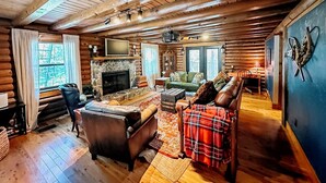 Een blokhutinterieur met een stenen open haard, houten balken in het plafond, een televisie en een gezellig zithoek met een geruit plaid.