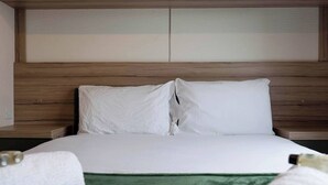 Un lit bien fait, avec du linge de lit blanc et un plaid vert, une tête de lit en bois et une table de chevet sur laquelle se trouvent une bouteille et une serviette.