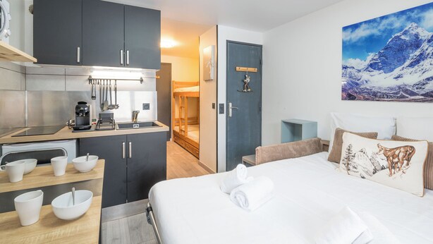A modern hotel room with a kitchenette, a bed with white linens, and a mountain landscape painting. - Studio L'étoile des neiges - Welkeys (Demi-Quartier)