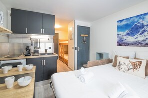 Une chambre d’hôtel moderne équipée d’une kitchenette, d’un lit avec du linge de maison blanc et d’un tableau représentant un paysage de montagne.