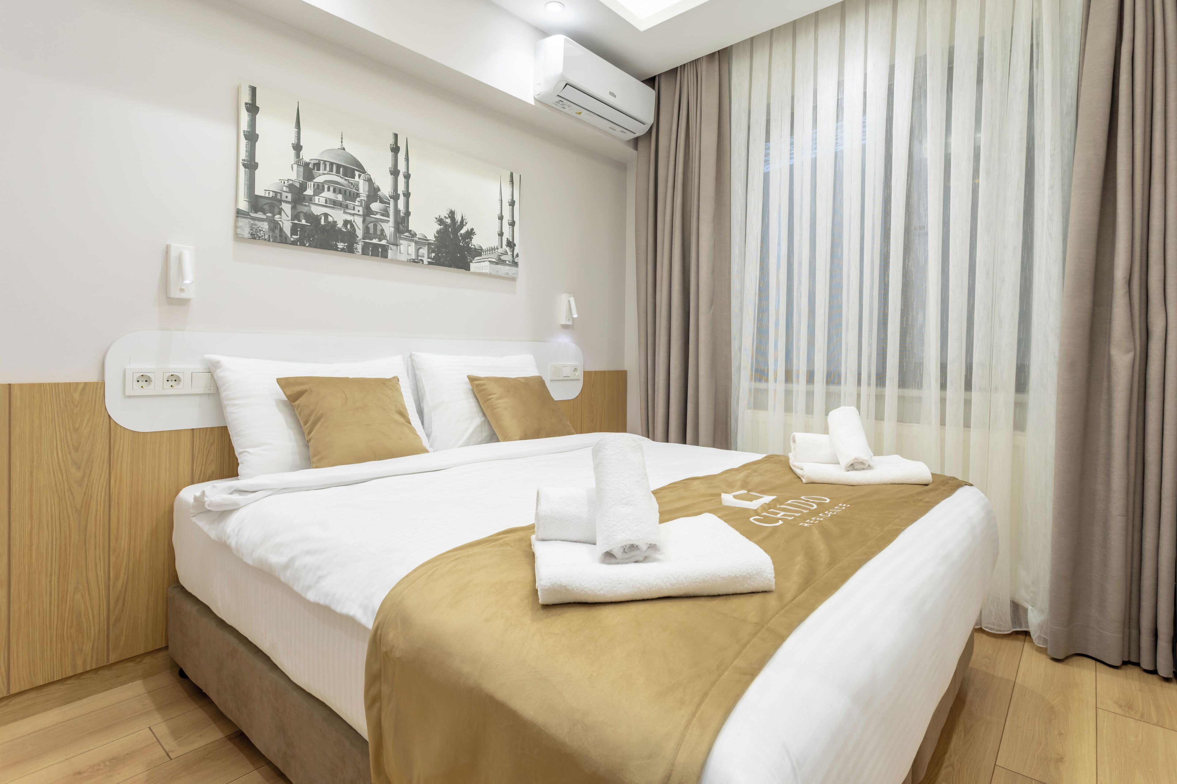 Ein Hotelzimmer mit einem ordentlich bezogenen Bett, einem Stadtbild als Wandkunst und einem Fenster mit Vorhängen.