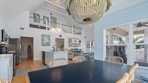 A spacious dining area with a long table and wicker chairs, a kitchen with white cabinets, and a large chandelier. - Pool - The Sea Pearl by SeaBreeze Vacation (Holmes Beach)