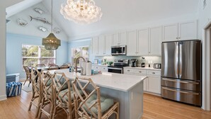 A kitchen with white cabinets, a stainless steel refrigerator, a large island with chairs, and a chandelier. - Pool - The Sea Pearl by SeaBreeze Vacation (Holmes Beach)