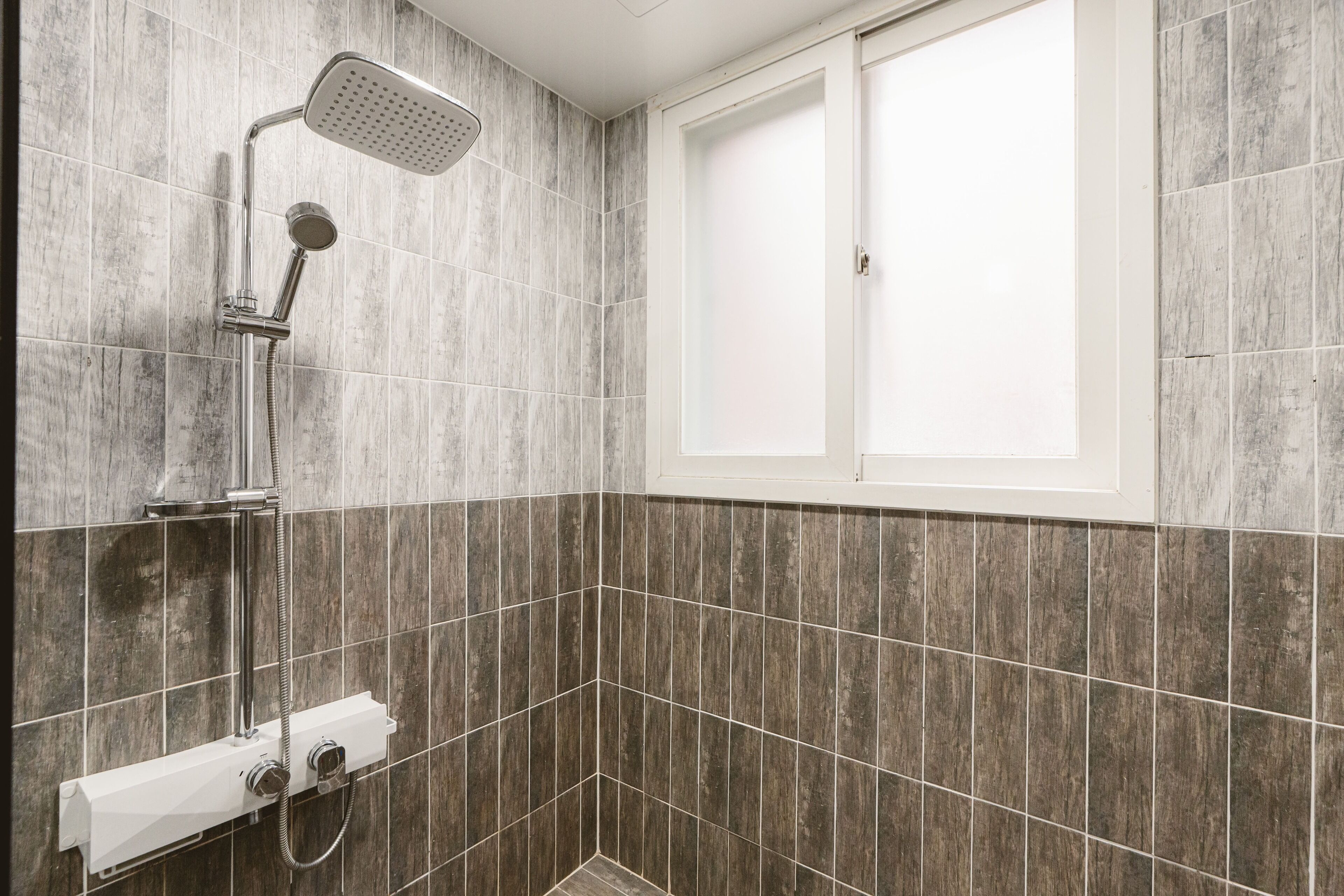 Un espace douche équipé d’un pommeau de douche à main, d’un panneau de commande fixé au mur et d’une fenêtre avec des stores fermés.
