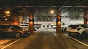 Garagem de estacionamento com vários carros, uma parede de tijolos de um lado e teto metálico com iluminação.