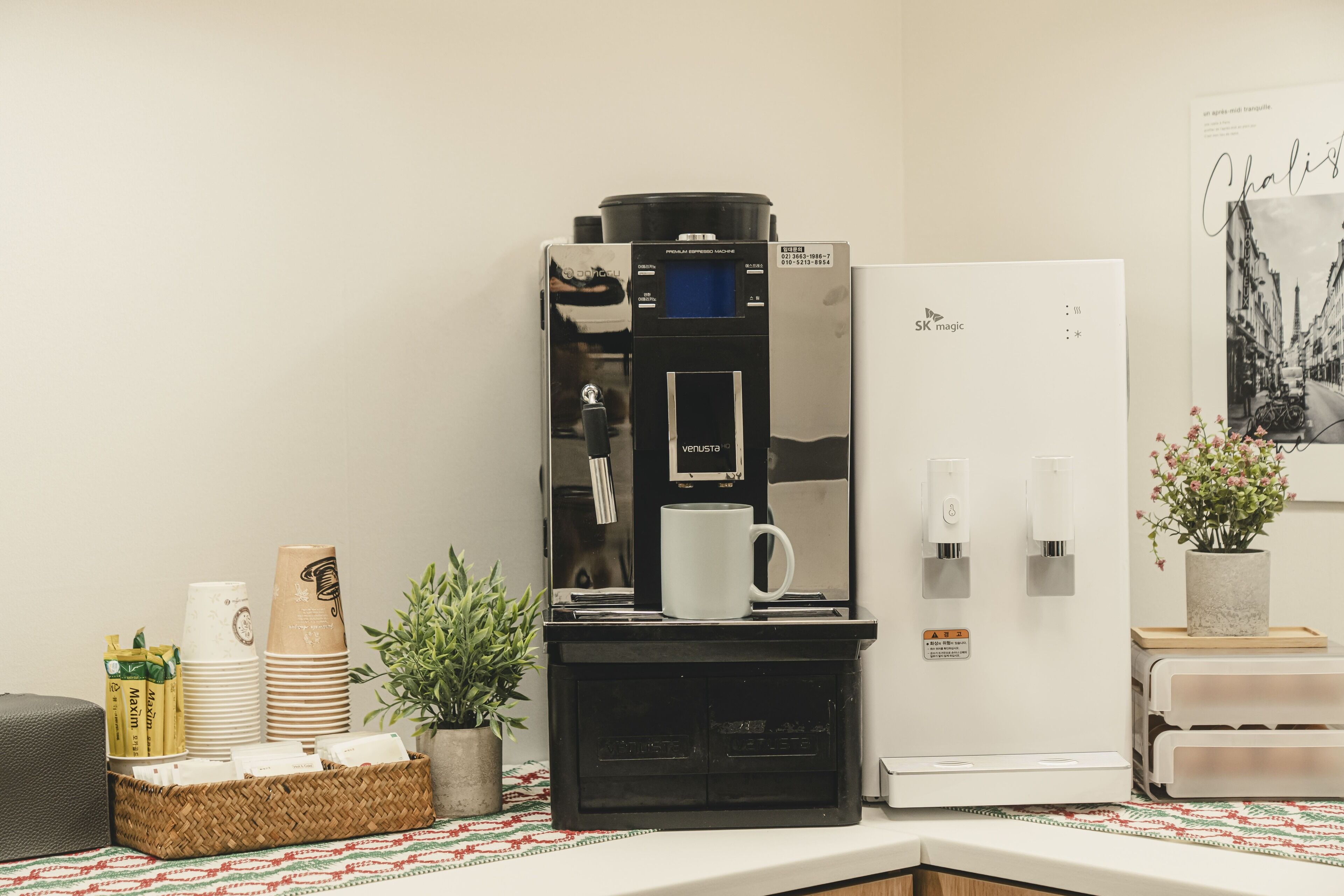 Et lille tekøkken med kaffemaskine, stablede papbægre, en kurv og en lille potteplante.