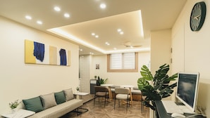 Uma área de espera moderna com sofá, cadeiras, uma mesa com computador e uma grande planta.