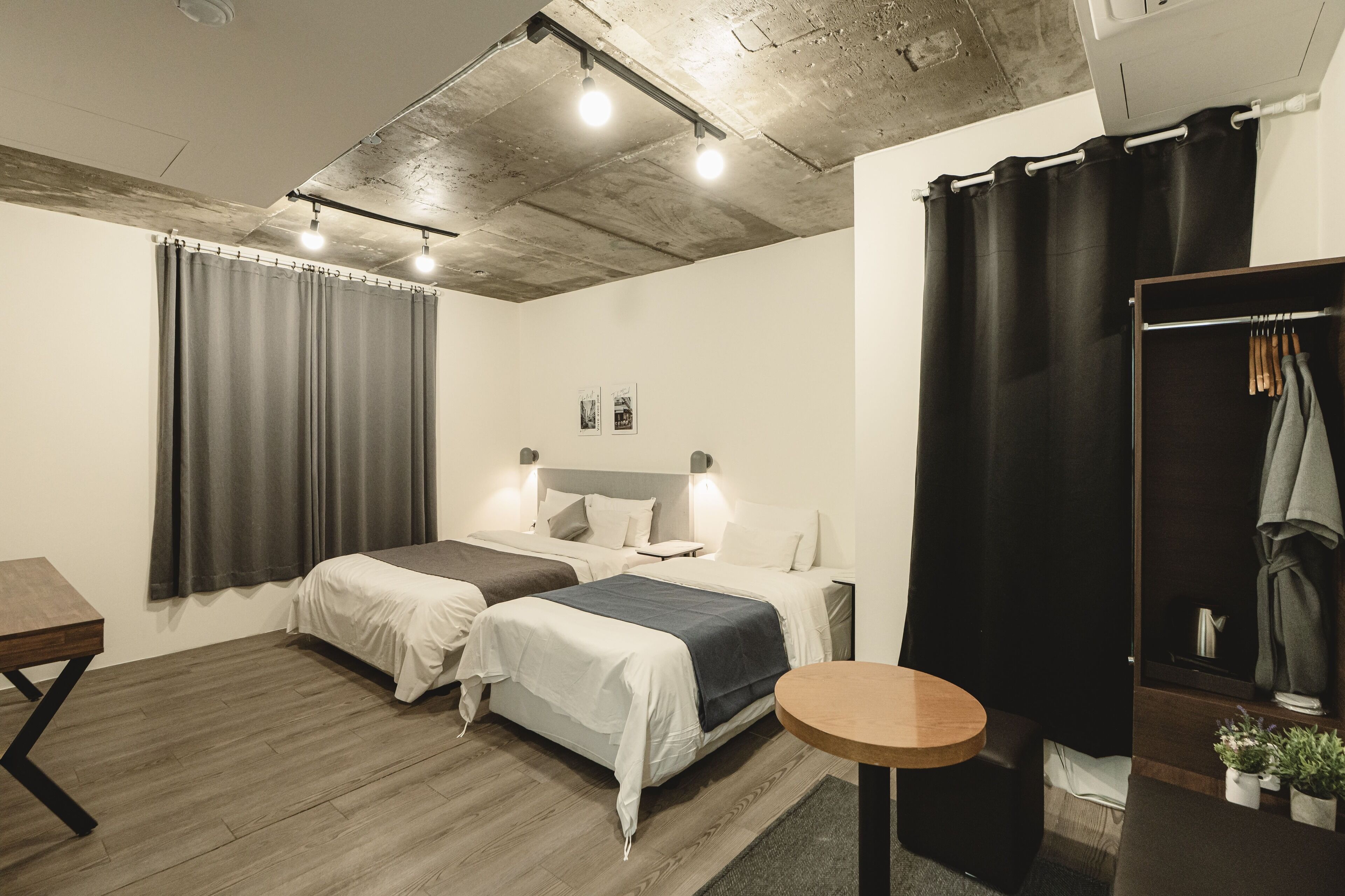 A modern hotel room with two beds, a small round table, a wardrobe, and a shower area.
