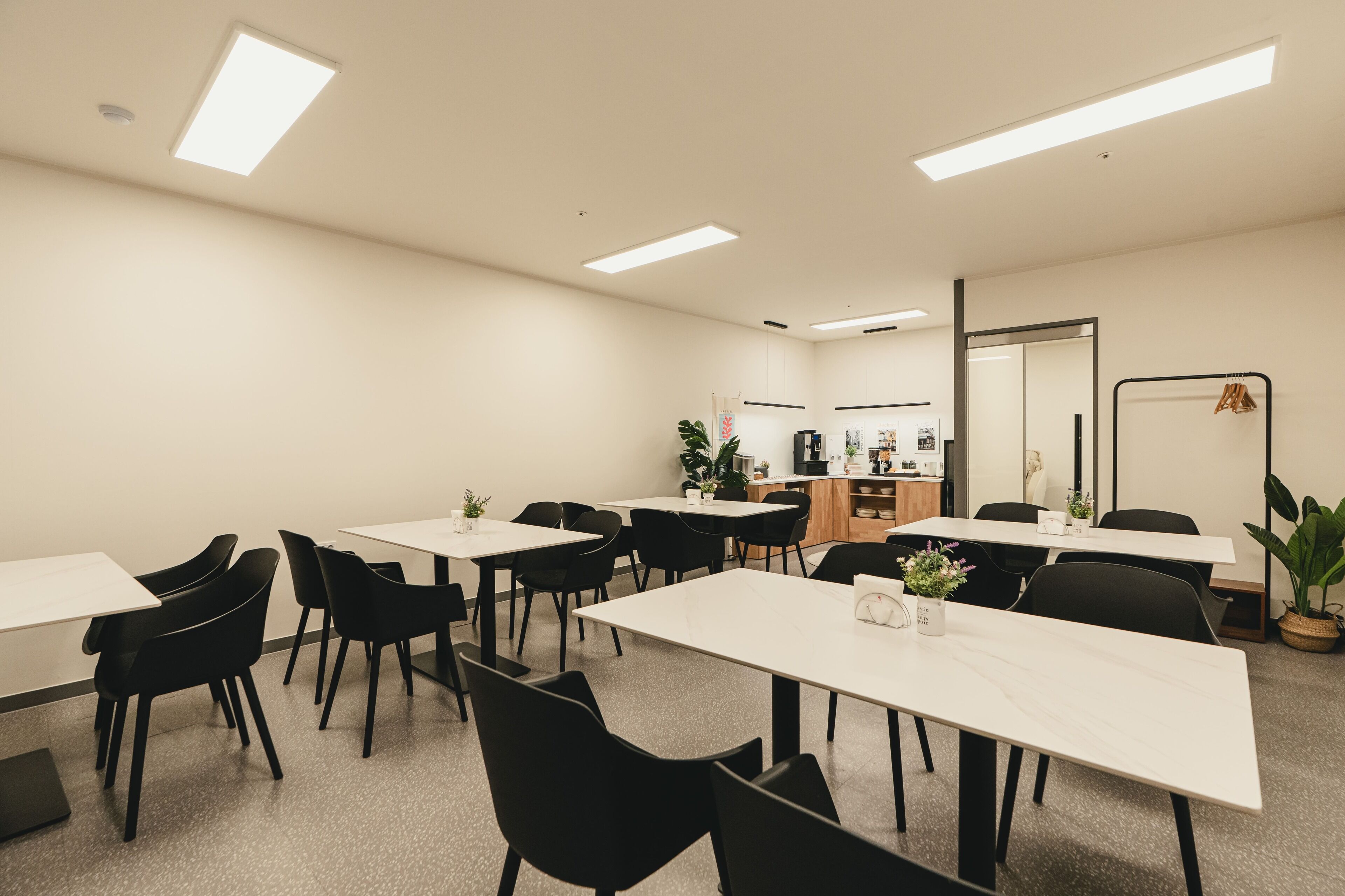 A conference room with white tables and black chairs, a kitchenette area, and a large mirror on the wall.
