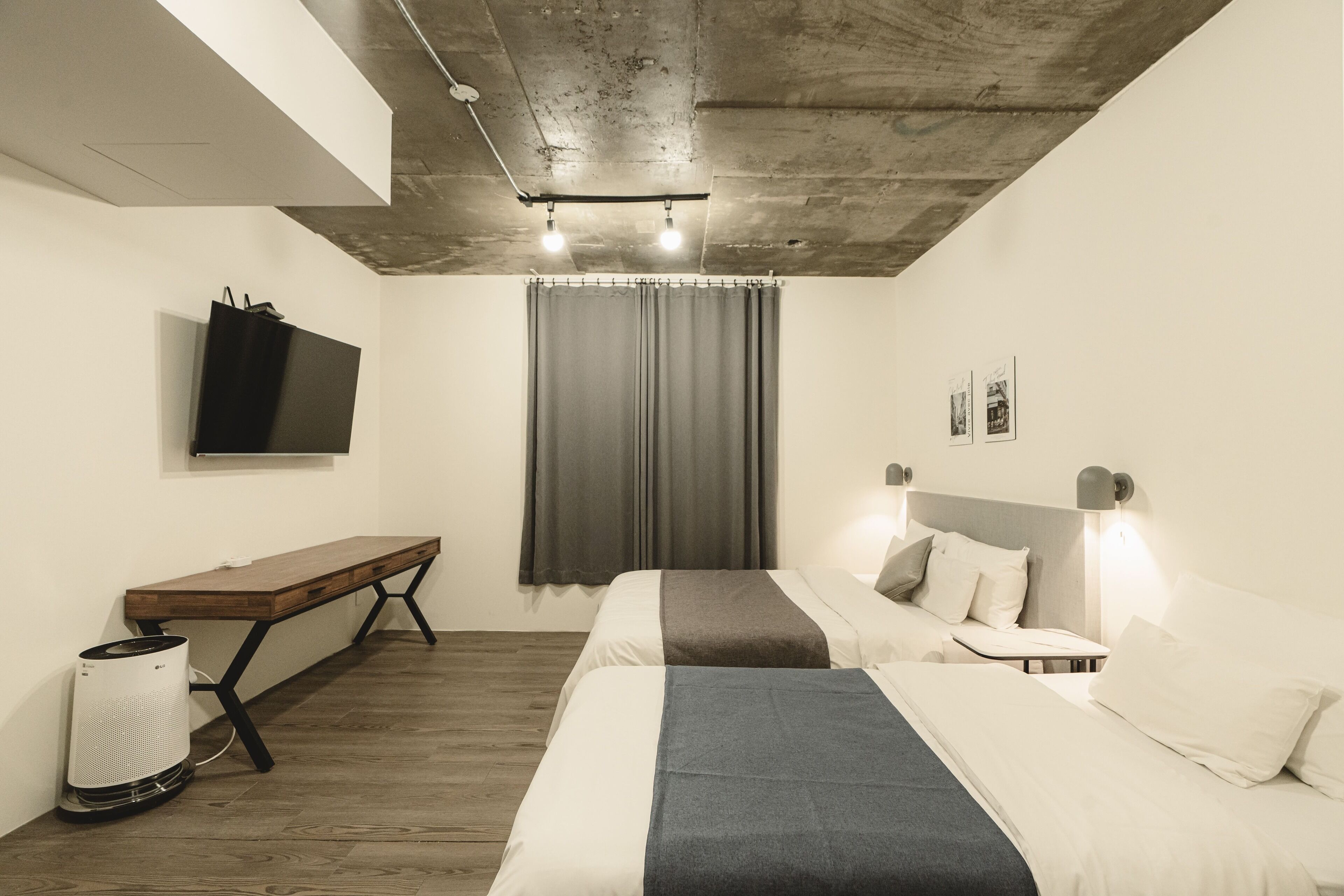 A modern hotel room with a wooden bench, a bed with a gray blanket, a small table, a wall-mounted TV, and a ceiling light fixture.