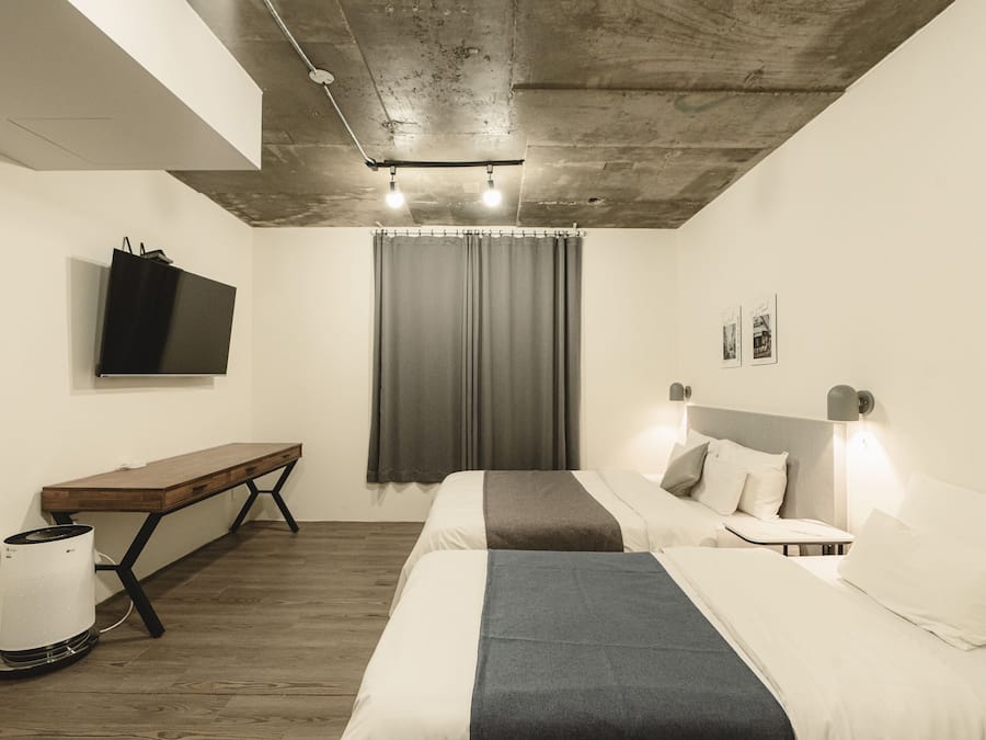 A modern hotel room with a wooden bench, a bed with a gray blanket, a small table, a wall-mounted TV, and a ceiling light fixture.