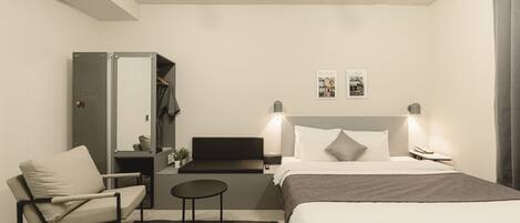 A modern hotel room with a bed, a chair, a small table, a mirror, and a nightstand.