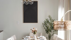 A dining area with a round table, chairs, a hanging light fixture, a wall-mounted artwork, and a plant.