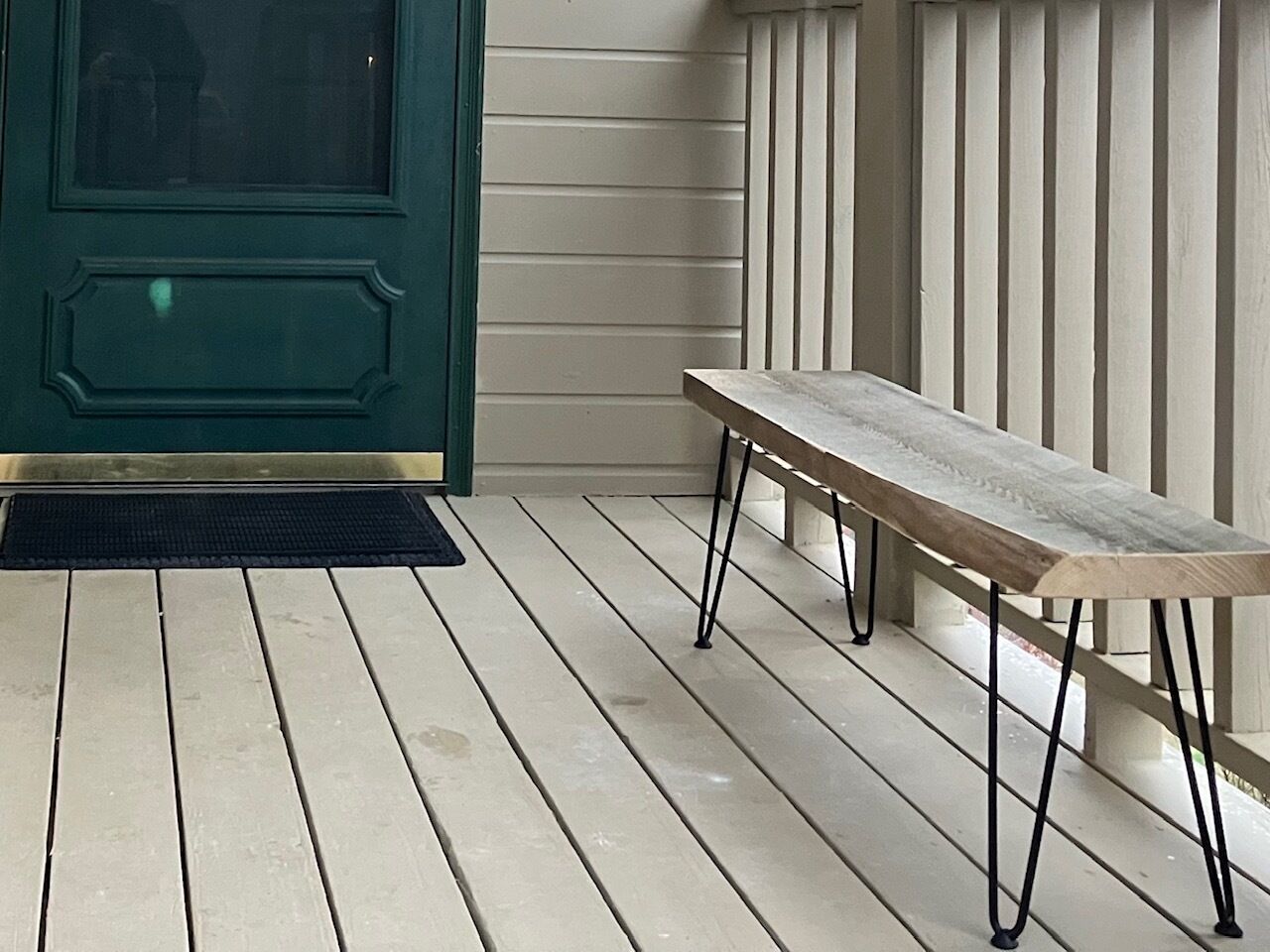Een houten bank met metalen poten op een veranda met witte houten planken en een groene deur.