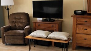 Une pièce comprenant un fauteuil marron, un téléviseur posé sur un meuble en bois, une petite table avec deux coussins, une lampe et une commode.