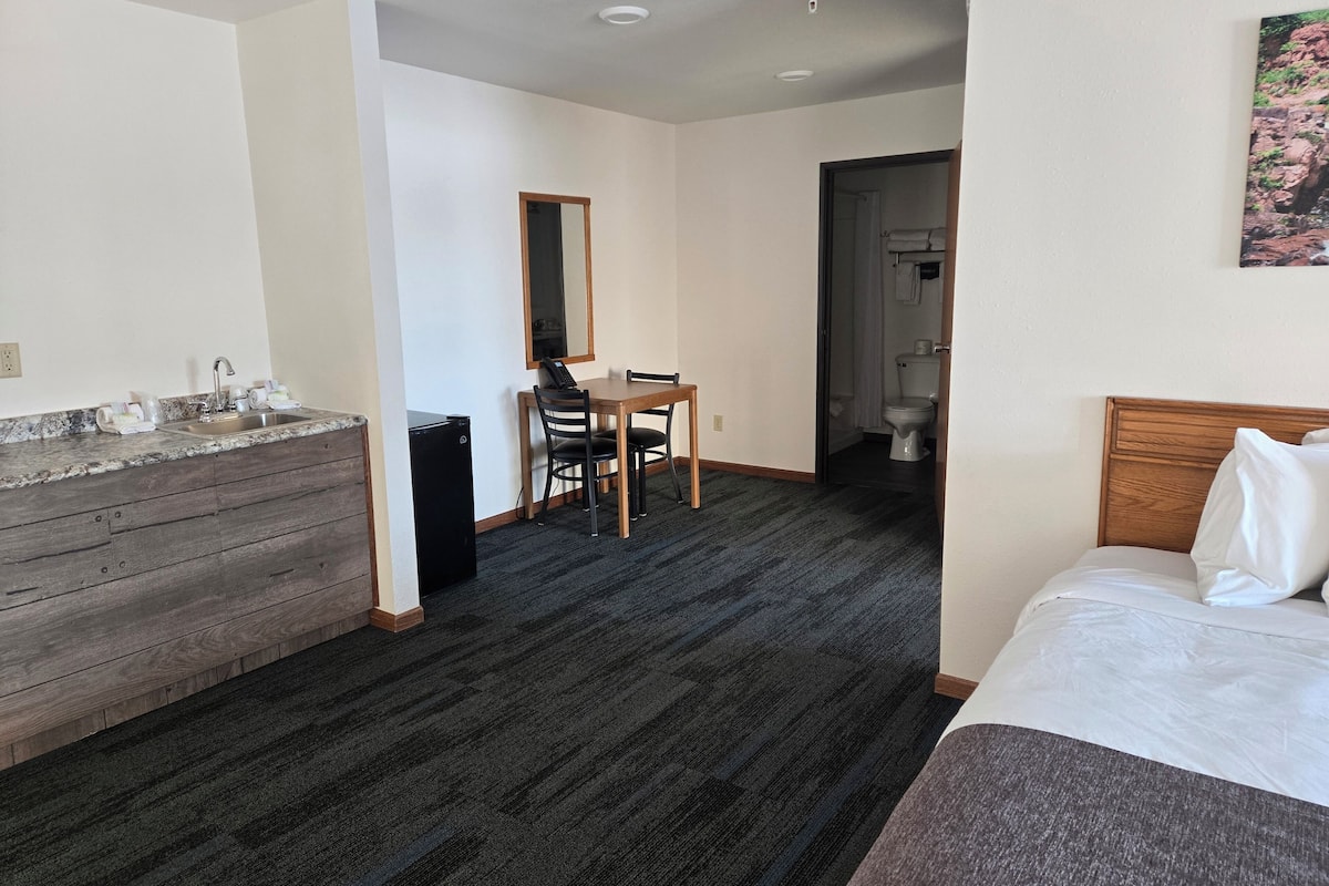 A hotel room with a bed, a desk with a chair, a small kitchenette, and a bathroom visible through an open door.