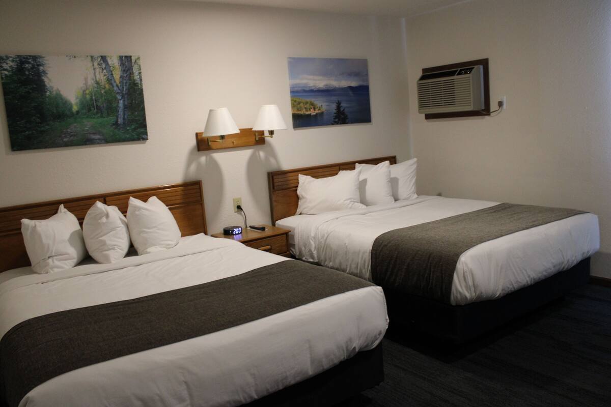 A hotel room with two beds, a nightstand, a wall-mounted air conditioner, and two paintings.
