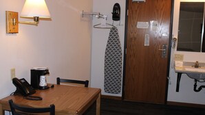 A hotel room with a wooden table, chairs, a coffee maker, a telephone, a wall-mounted lamp, an ironing board, a mirror, and a sink.