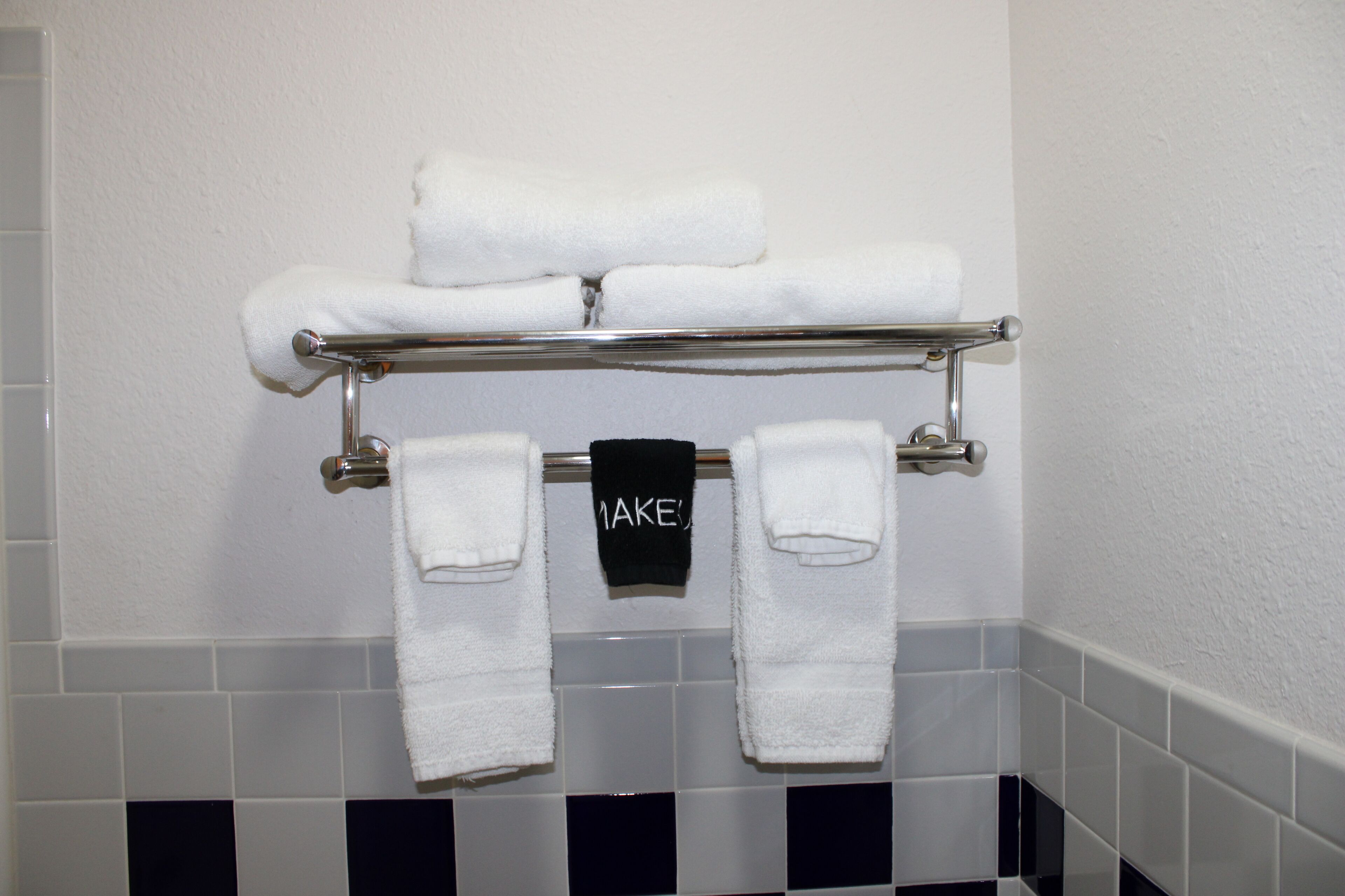 A bathroom with a towel rack holding three white towels and a black towel with the word "MAKES" on it.