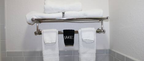 Un baño con un perchero que sostiene tres toallas blancas y una toalla negra con la palabra 'MAKES' escrita.