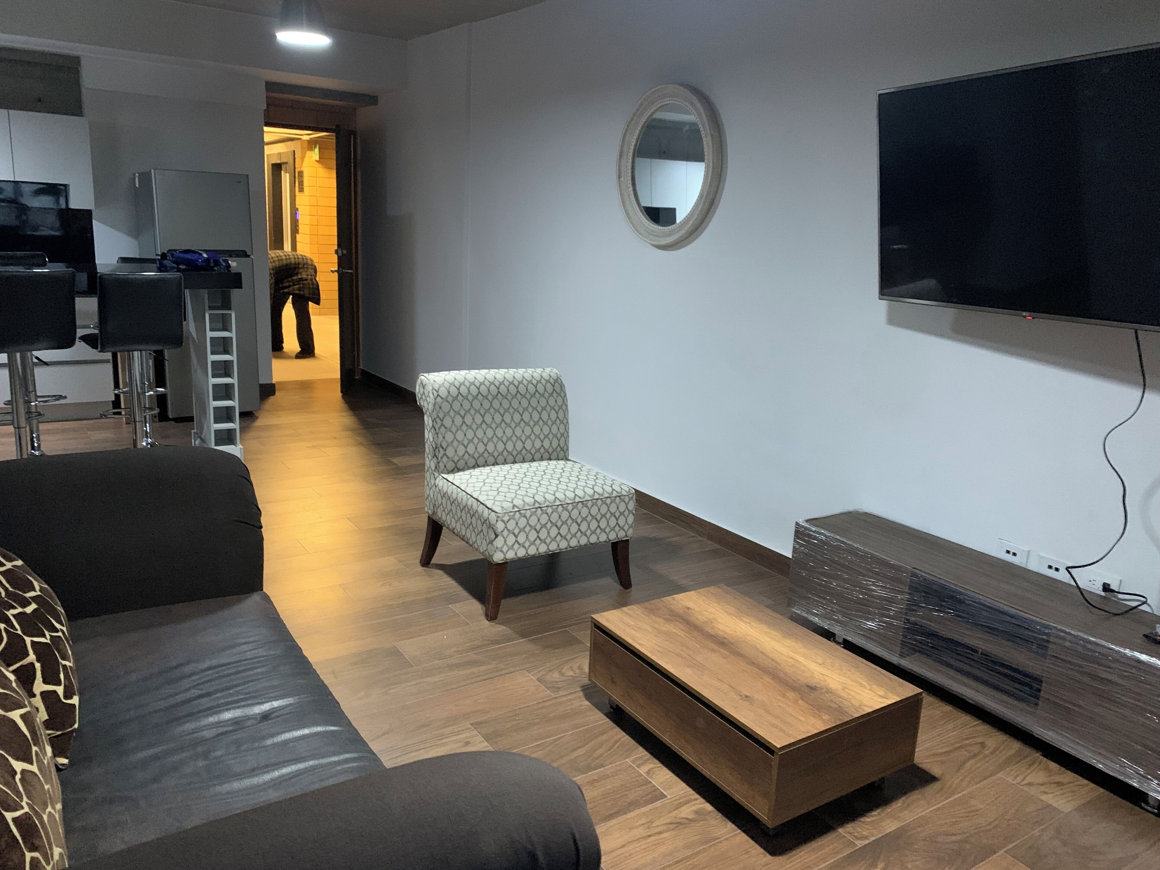 Una sala moderna con un sofá, un sillón estampado, una mesa de centro de madera, un televisor de pantalla plana y un espejo en la pared.