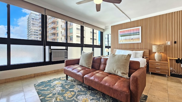 A hotel room with a large window, a brown leather sofa, a bed, a wooden wall panel, and a painting. - Waikiki Grand Hotel® 521—Honolulu Ocean side Studio Sleeps 3 w/ Kitchen & Pool (Honolulu)