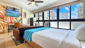 A hotel room with a large bed, a desk with a chair, a ceiling fan, and a city view through large windows. - Waikiki Grand Hotel® 521—Honolulu Ocean side Studio Sleeps 3 w/ Kitchen & Pool (Honolulu)