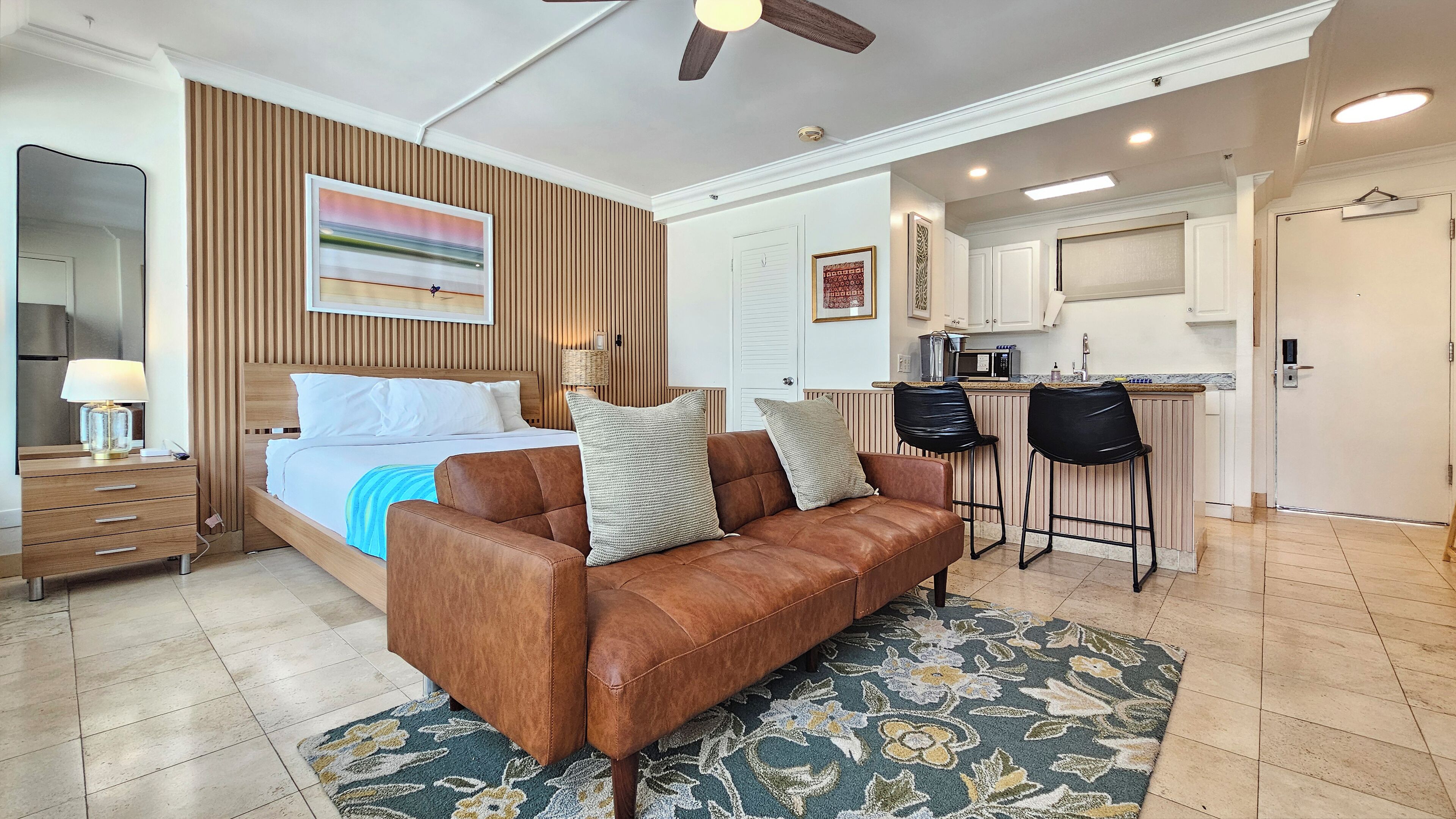 A modern hotel room with a bed, a brown leather sofa, a dining area with a bar, and a kitchen.