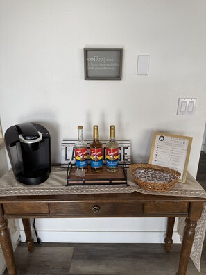 Eine Kaffeemaschine, drei Flaschen Torani-Sirup, ein gerahmtes Willkommensschild und eine Korb mit Kaffeekapseln auf einer Holztisch.